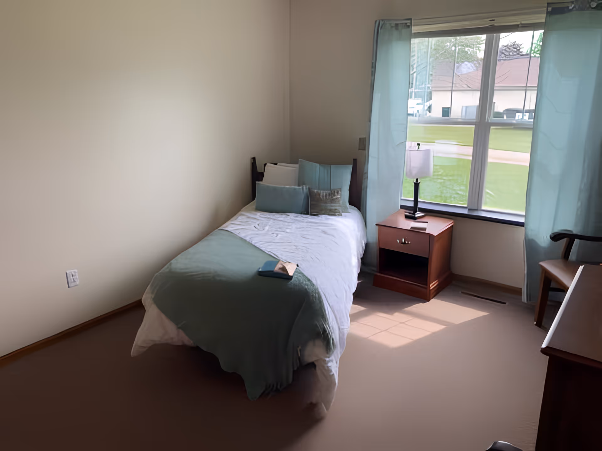 A small, neatly arranged bedroom with a single bed covered in white and light green bedding. There are several pillows on the bed. Next to the bed is a wooden nightstand with a lamp and a book. A large window with light blue curtains lets in natural light, showing a green lawn outside. A wooden chair and part of a desk are visible on the right side of the room.