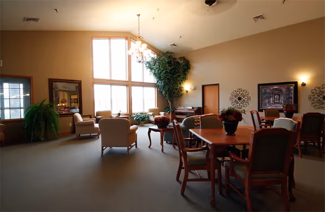 A spacious, well-lit common area with large windows allowing natural light to fill the room. The room features several upholstered armchairs arranged around small tables, a chandelier hanging from the ceiling, decorative plants, wall art, and a piano against the far wall.