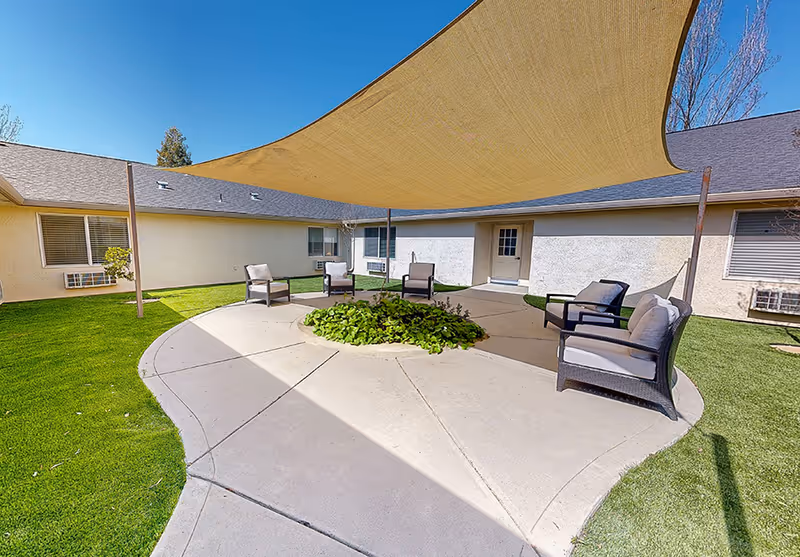 Outdoor courtyard area with a circular concrete patio surrounded by green grass. The patio has a central planter with green plants and is shaded by a large beige fabric canopy. Several cushioned chairs are arranged around the patio. The surrounding building has light-colored walls and windows with air conditioning units.