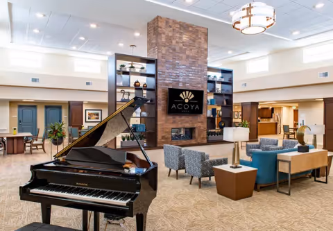 Spacious and well-lit living room area in ACOYA Mesa featuring a black grand piano in the foreground, a central stone fireplace with a mounted TV displaying the ACOYA Mesa logo, modern seating arrangements including blue and patterned chairs, decorative shelves, and a high ceiling with large windows allowing natural light.