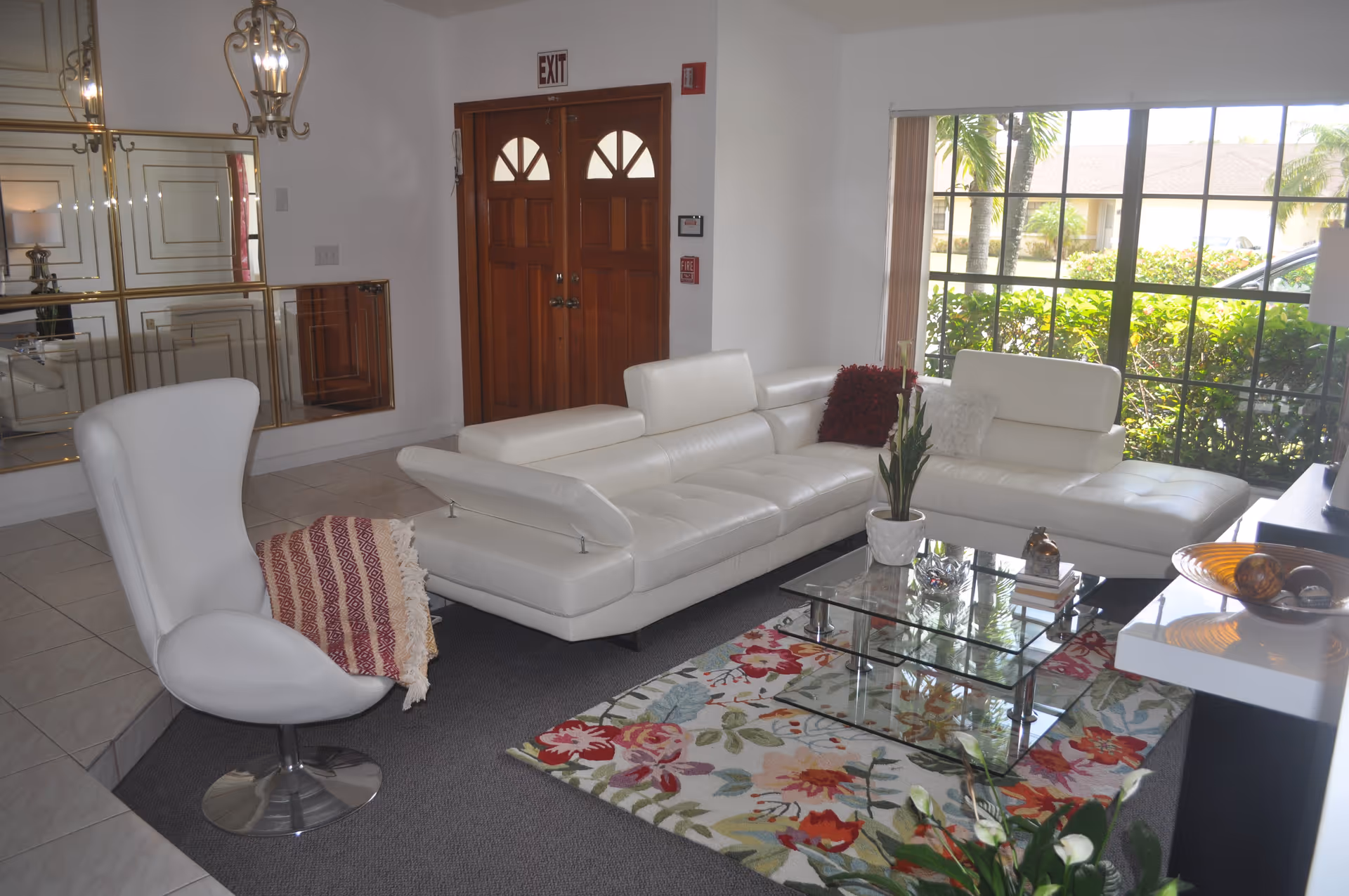 A bright living room with a white modern sectional sofa and a white swivel chair with a red and white patterned throw. There is a glass coffee table on a floral rug, a large window showing greenery outside, and a wooden double door with an exit sign above it. The room has mirrored wall panels and a chandelier.