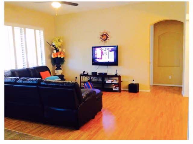 A living room with a black leather sectional sofa, a flat-screen TV mounted on a beige wall, a wooden TV stand with various electronic devices, a decorative sun wall hanging above the TV, a large vase with flowers in the corner, and wooden flooring.