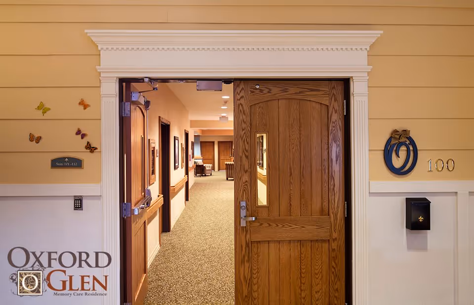 Open wooden door leading into a carpeted interior hallway of a memory care facility with the number 100 visible and the Oxford Glen logo.