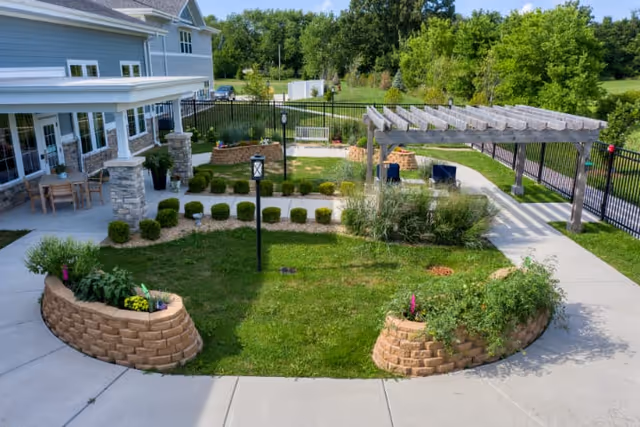 Outdoor garden area at Cedarhurst of Yorkville featuring a green lawn, raised brick flower beds with plants, a pergola with two chairs underneath, a paved walkway, and a building with a covered patio and outdoor seating on the left side. The area is enclosed by a black metal fence and surrounded by trees and greenery.