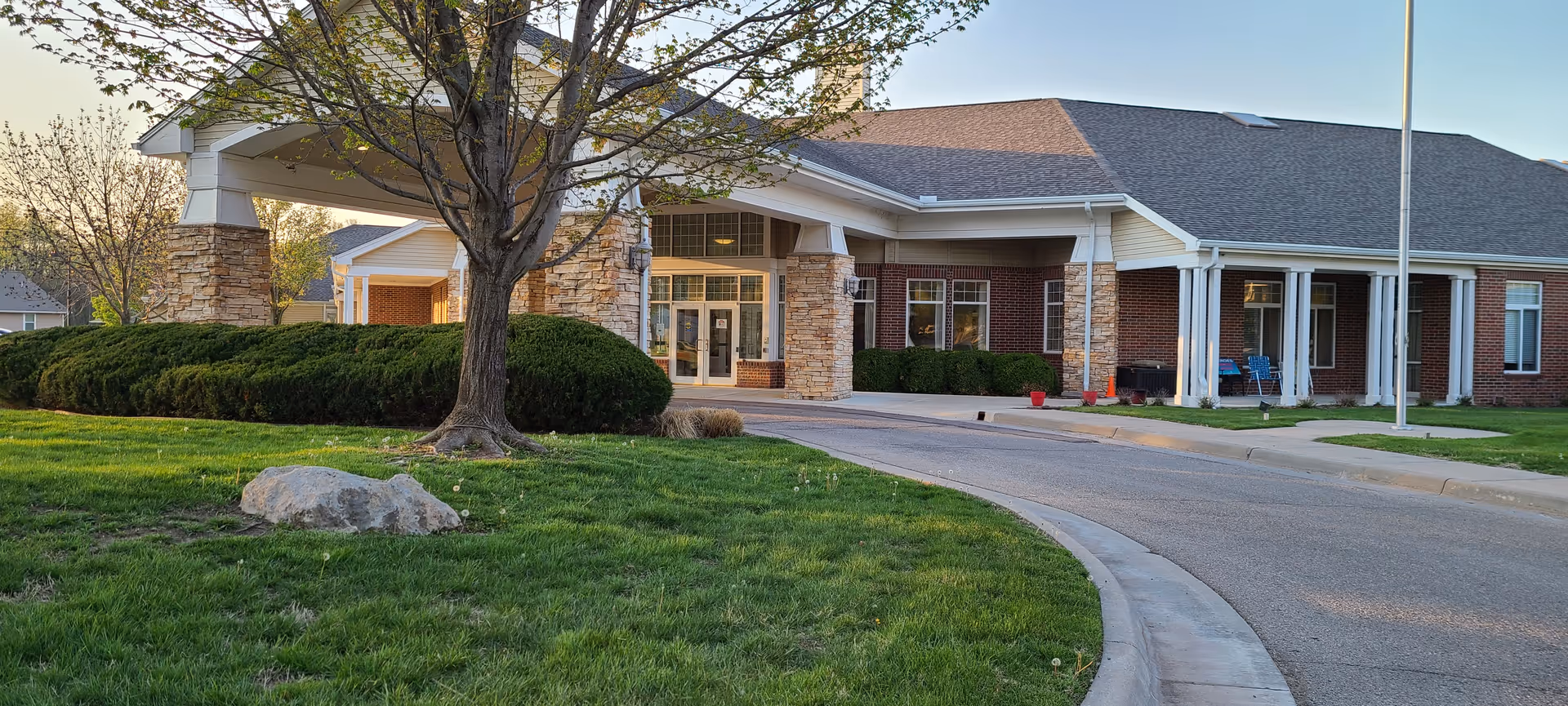 Front entrance of a single-story brick senior living building with a porte-cochere, landscaped lawn, and curved driveway.
