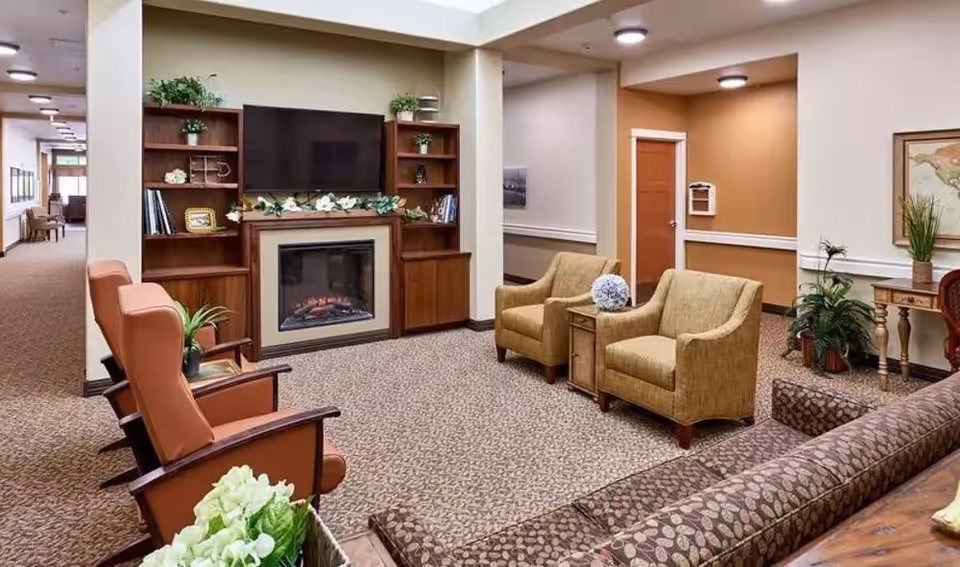 A cozy living room area in a senior living facility featuring a wall-mounted flat-screen TV above an electric fireplace, flanked by wooden shelves with decorative plants and books. The room has two beige armchairs with a small wooden side table between them, two brown cushioned chairs, and a patterned sofa. The walls are painted in neutral tones with a mix of white and light brown, and there are plants and framed artwork decorating the space.