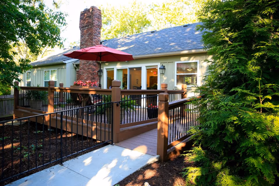 A wooden wheelchair-accessible deck with railings and a red patio umbrella attached to a single-story house surrounded by trees.