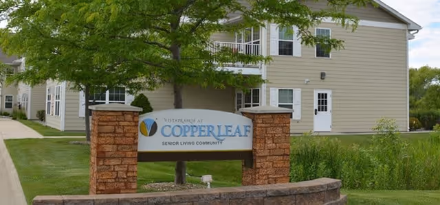 A stone-framed sign reading "Vista Prairie at Copperleaf Senior Living Community" sits in front of a beige two-story residential building with trees and landscaping.