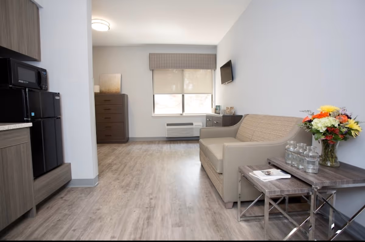 A modern living area in a senior living facility with light wood flooring, a beige sofa, a side table with a vase of colorful flowers and glass jars, a window with a blind, a wall-mounted TV, and kitchen appliances including a microwave and mini refrigerator.