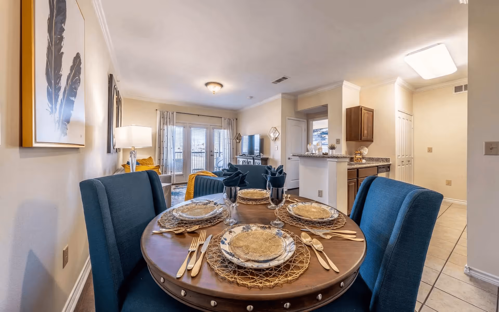 A dining area with a round wooden table set for four with plates, gold-colored cutlery, and dark blue napkins in wine glasses. The dining chairs are upholstered in blue fabric. In the background, there is a living room with a sofa, a TV on a stand, and glass doors with curtains leading outside. To the right, there is a kitchen with wooden cabinets and granite countertops.