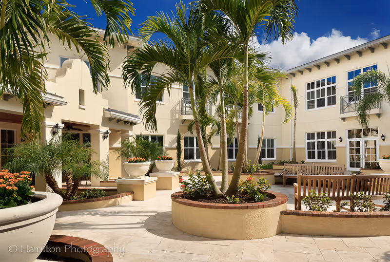 Sunny courtyard with palm trees, large planters, wooden benches and a two-story cream-colored building under a blue sky.