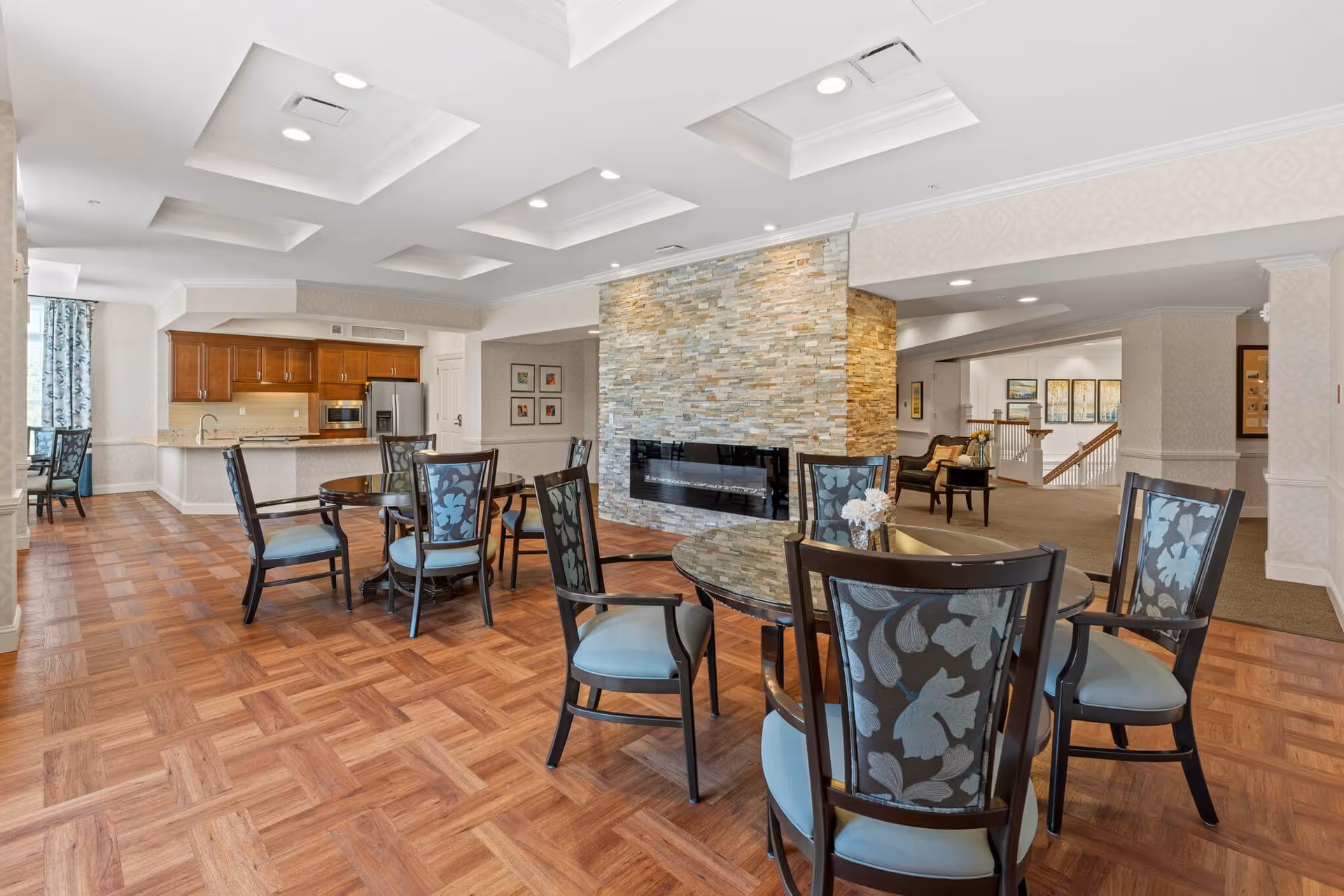 Open communal dining and lounge area with round tables and chairs, a stone fireplace divider, and a kitchen in the background.