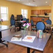 Bright communal dining/activity room with tables and chairs, a kitchenette along the back wall, and flowers and magazines on the nearest table.