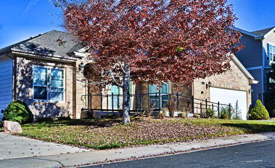 Exterior view of a single-story brick residential building with a large tree with red leaves in front. The building has multiple windows with blinds and a garage door. There is a concrete sidewalk and a ramp with black railings leading to the entrance. The lawn has some fallen leaves and small bushes.