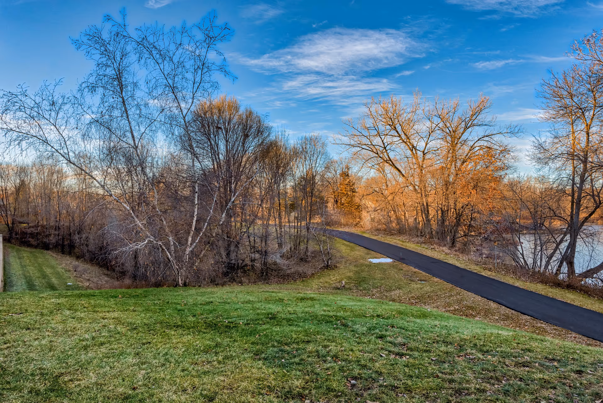 A scenic outdoor view featuring a paved walking path winding through a grassy area with leafless trees and a body of water visible on the right side under a partly cloudy blue sky.
