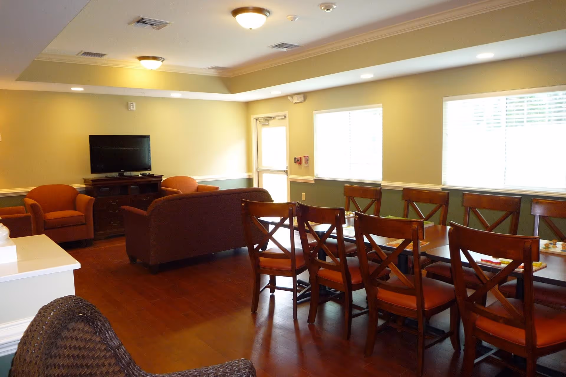 A well-lit room with a dining area featuring a long wooden table surrounded by wooden chairs with orange cushions. In the background, there is a seating area with a brown couch and two orange armchairs facing a flat-screen TV on a wooden stand. The room has light green walls, large windows with blinds, and wooden flooring.