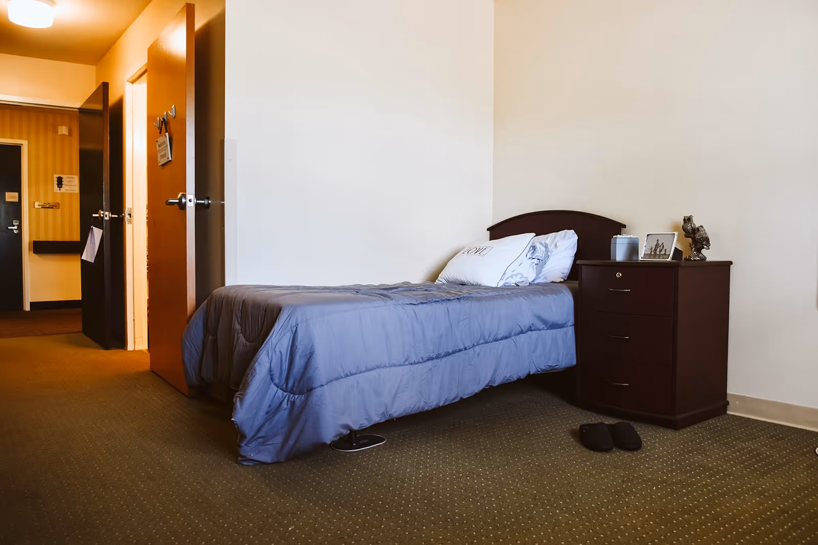 A simple bedroom in a senior living facility with a single bed covered in a blue comforter, a pillow with the word 'LOVE' on it, a dark wooden nightstand with decorative items including a framed photo and a small owl figurine, and a pair of black slippers on the carpeted floor. The room has beige walls and an open door leading to a hallway.
