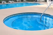 A circular hot tub with clear blue water and a metal handrail, adjacent to a larger rectangular swimming pool surrounded by white lounge chairs.
