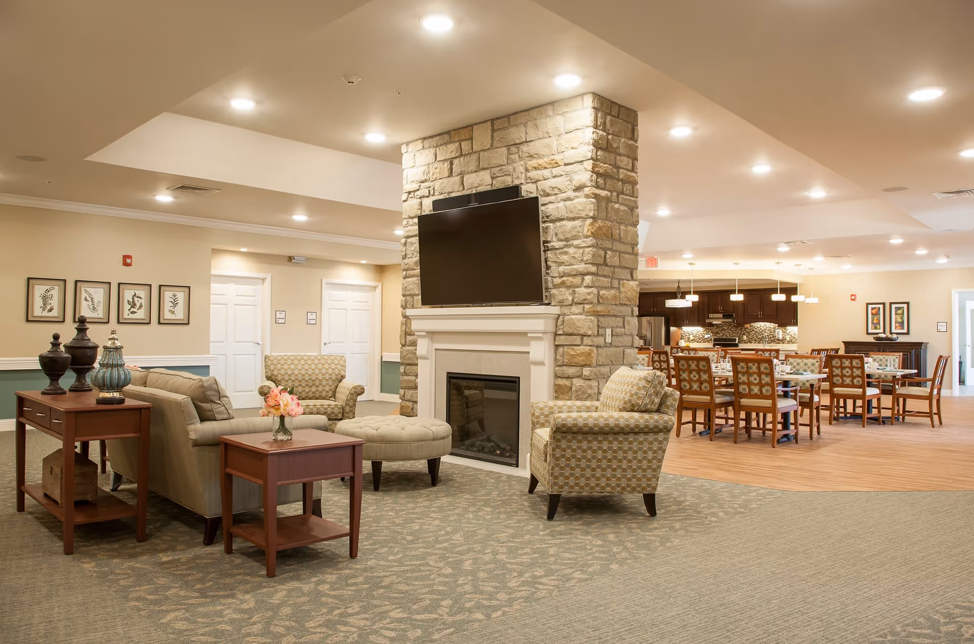 A spacious senior living facility common area featuring a stone fireplace with a mounted flat-screen TV above it. The seating area includes a sofa, armchairs, and an ottoman arranged around the fireplace. In the background, there is a dining area with multiple tables and chairs, and a kitchen with dark wood cabinets and pendant lighting. The room is well-lit with recessed ceiling lights and decorated with framed artwork on the walls.