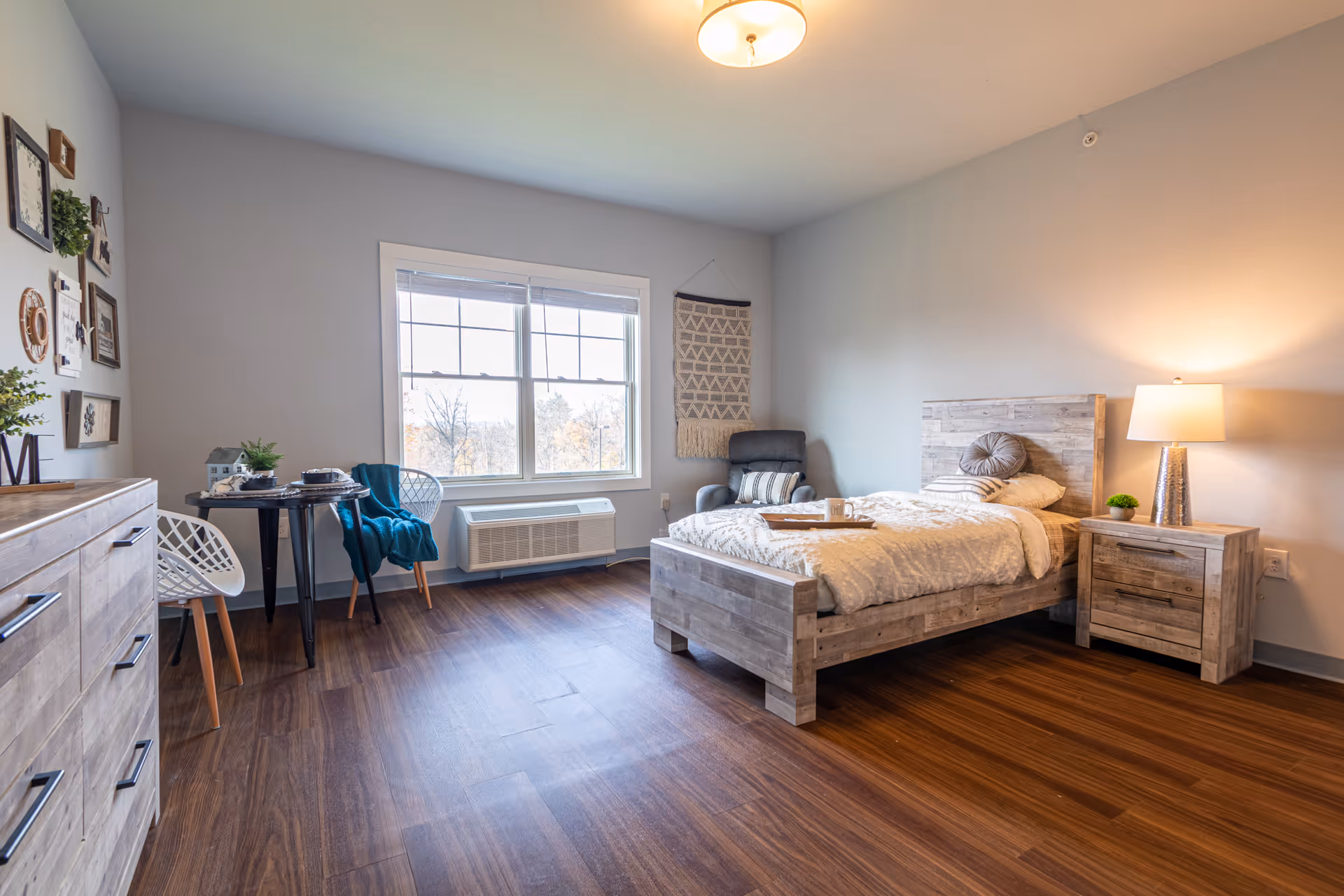 A bright and spacious bedroom with wooden flooring, a wooden bed with white bedding, a matching nightstand with a lamp, a comfortable armchair, a small round table with two chairs near a large window, and decorative wall art.