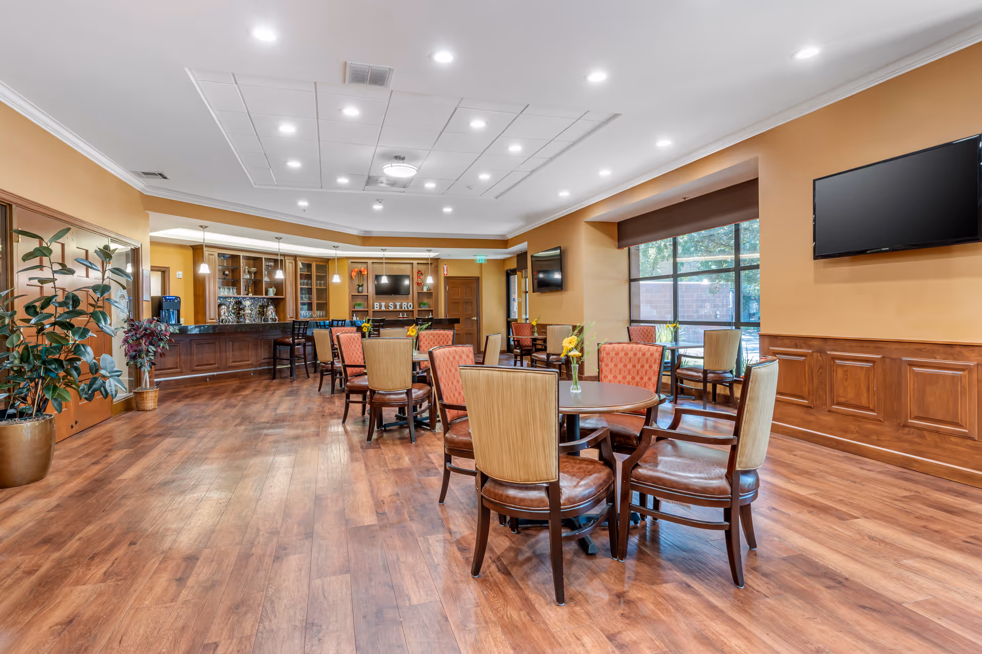 A well-lit dining area with wooden floors, multiple round tables surrounded by chairs with beige and red upholstery, a bar counter with high stools, large windows letting in natural light, and two wall-mounted flat-screen TVs.