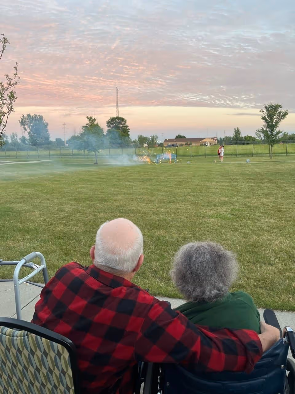 An elderly couple sitting outdoors on chairs, with the man wearing a red and black checkered shirt and the woman with gray hair, watching fireworks or sparklers on a grassy field during sunset. A walker is visible next to the man, and a person is seen in the distance near a fence.