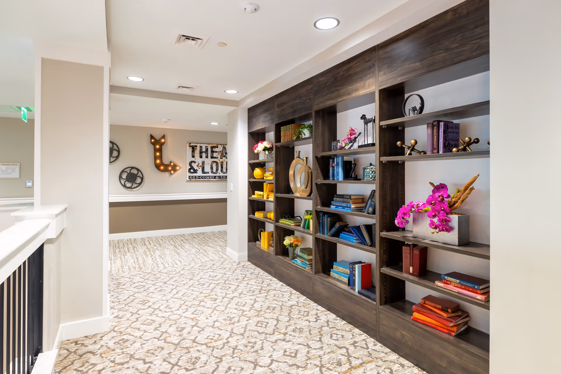 Bright interior hallway with a long built-in bookshelf filled with books and decorative items, patterned carpet, and wall decor.