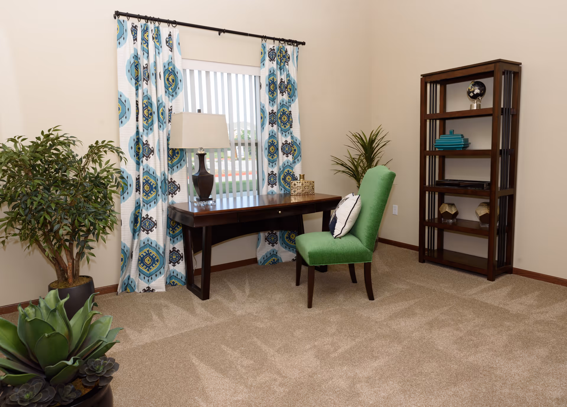 A cozy room with beige walls and carpet featuring a dark wooden desk with a lamp and decorative box on top. A green upholstered chair with a white pillow is placed in front of the desk. The window behind the desk has vertical blinds and blue patterned curtains. There are two potted plants in the room, one large leafy plant on the left and a smaller plant near a tall wooden bookshelf with decorative items on the right.