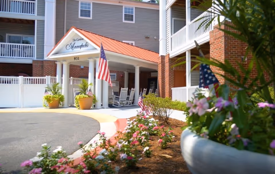 Entrance of The Gardens of Annapolis senior living facility with a covered porch supported by white columns, rocking chairs, American flags, potted plants, and a flower bed in the foreground.