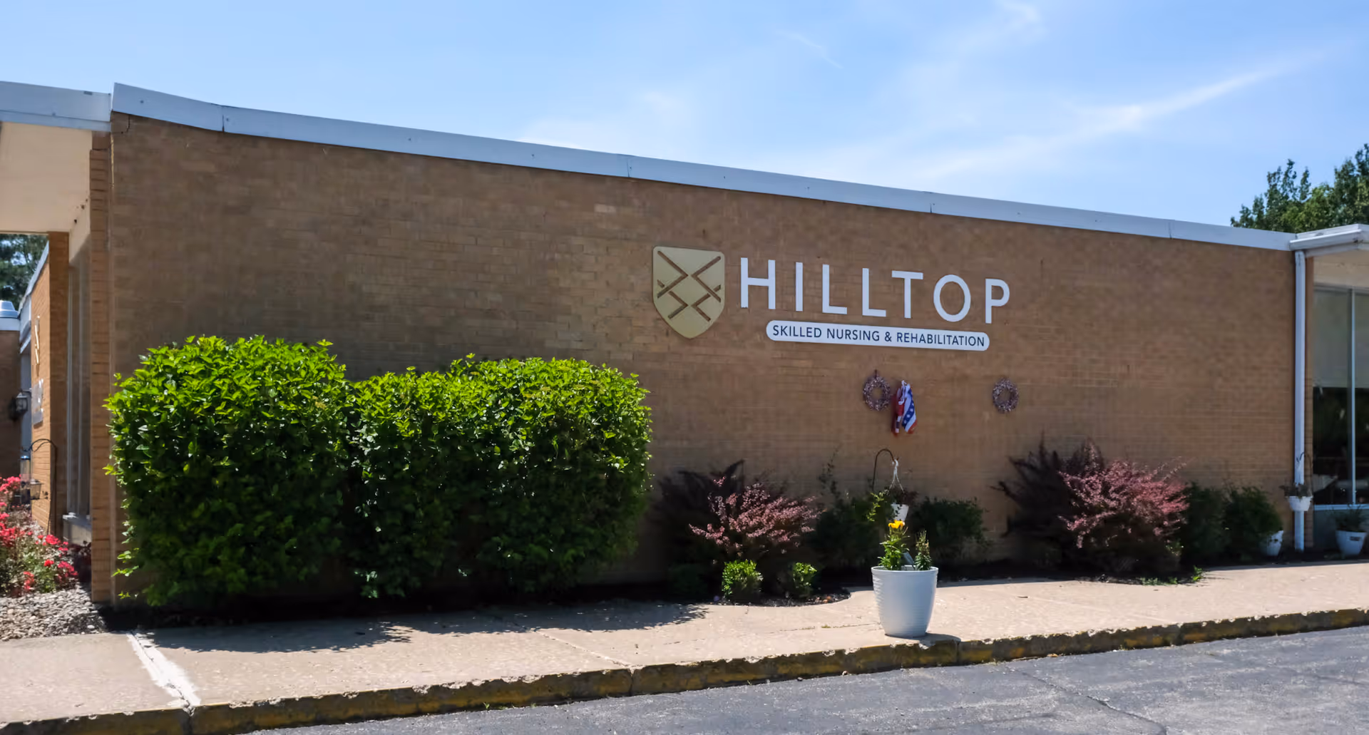 Exterior view of Hilltop Skilled Nursing and Rehabilitation building with a brick facade, green bushes, and a small potted plant near the sidewalk under a blue sky.