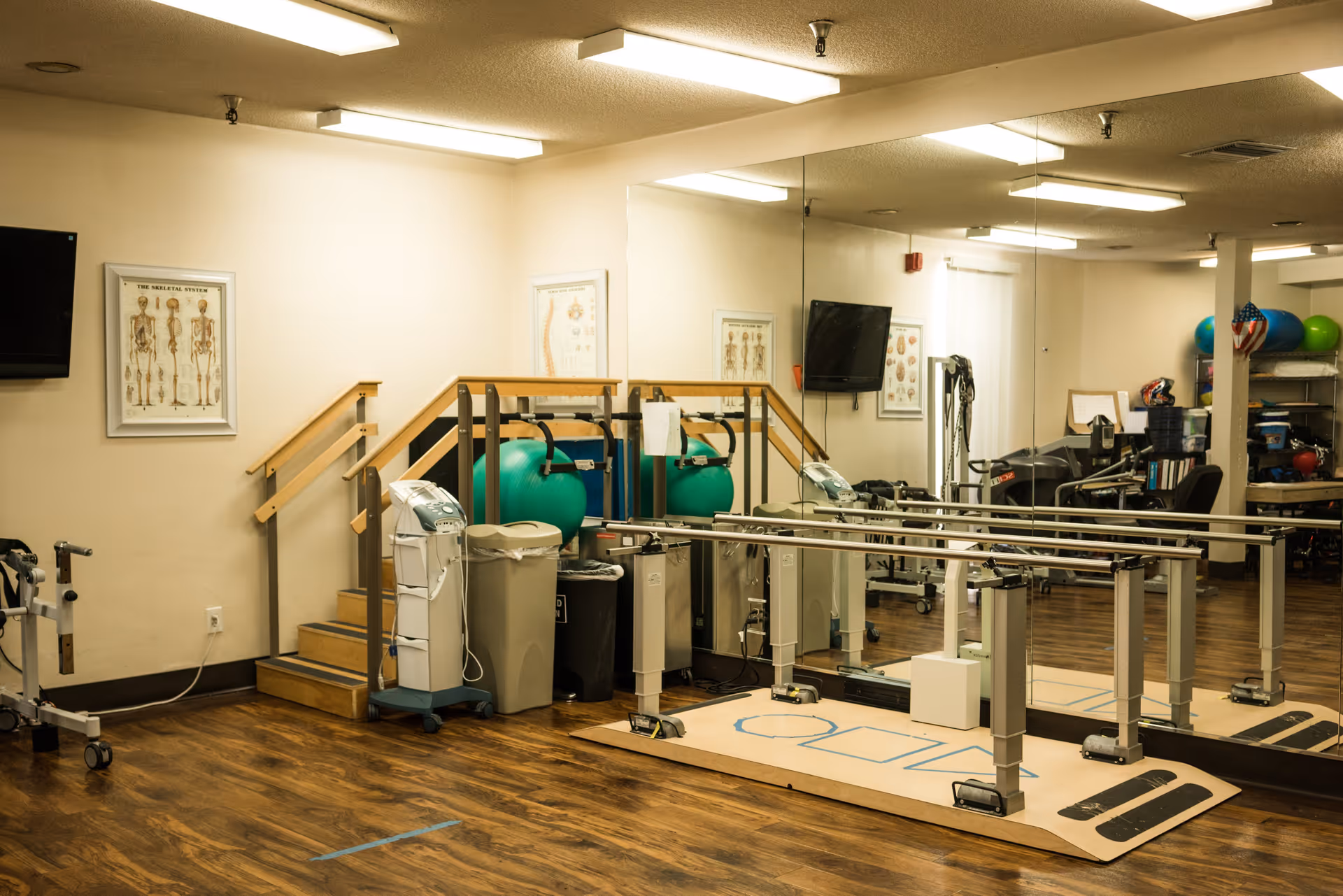 A physical therapy or rehabilitation room with wooden flooring, parallel bars for walking exercises, a small staircase with handrails, exercise balls, medical equipment, a wall-mounted TV, anatomical posters, and a large mirror covering one wall.