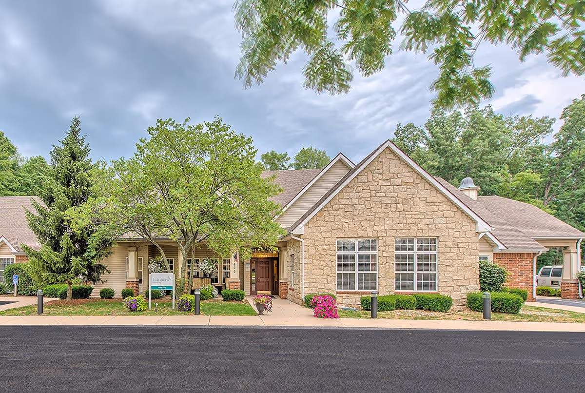 Front exterior view of Trail Creek Place Assisted Living facility with stone and brick facade, a tree and flower pots near the entrance, and a paved driveway in front.