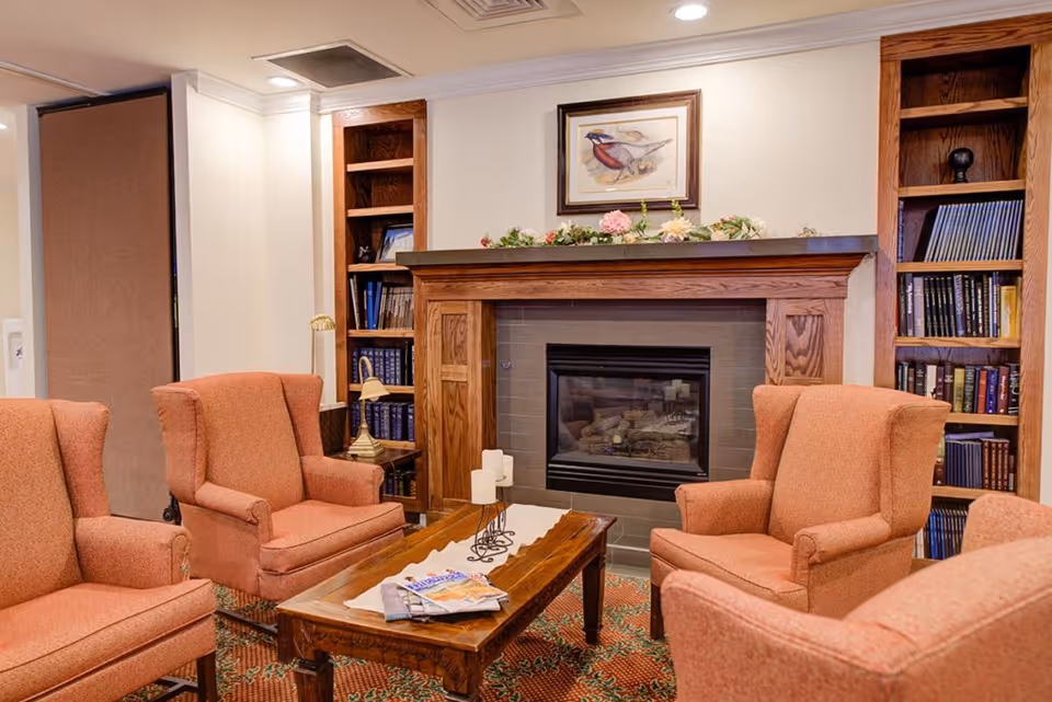 A cozy living room area with four orange upholstered armchairs arranged around a wooden coffee table. The coffee table has a white runner, candles, and magazines on it. Behind the chairs is a fireplace with a wooden mantel decorated with flowers and a framed bird painting above it. There are built-in wooden bookshelves on either side of the fireplace filled with books and decorative items.