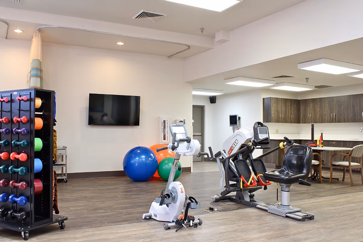 A rehabilitation exercise room with various fitness equipment including a stationary bike, a recumbent exercise machine, colorful dumbbells on a rack, and large exercise balls. The room has wood flooring, white walls, a wall-mounted TV, and a seating area with chairs and a table in the background.