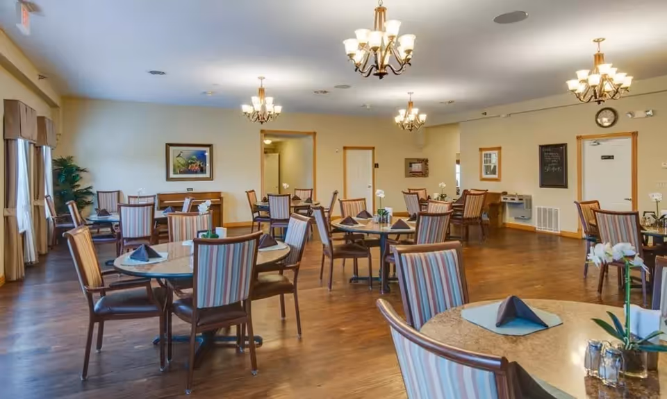 A spacious dining room with multiple round tables and striped cushioned chairs. Each table is set with napkins and small flower arrangements. The room has wooden flooring, beige walls, several chandeliers hanging from the ceiling, and large windows with curtains allowing natural light to enter.