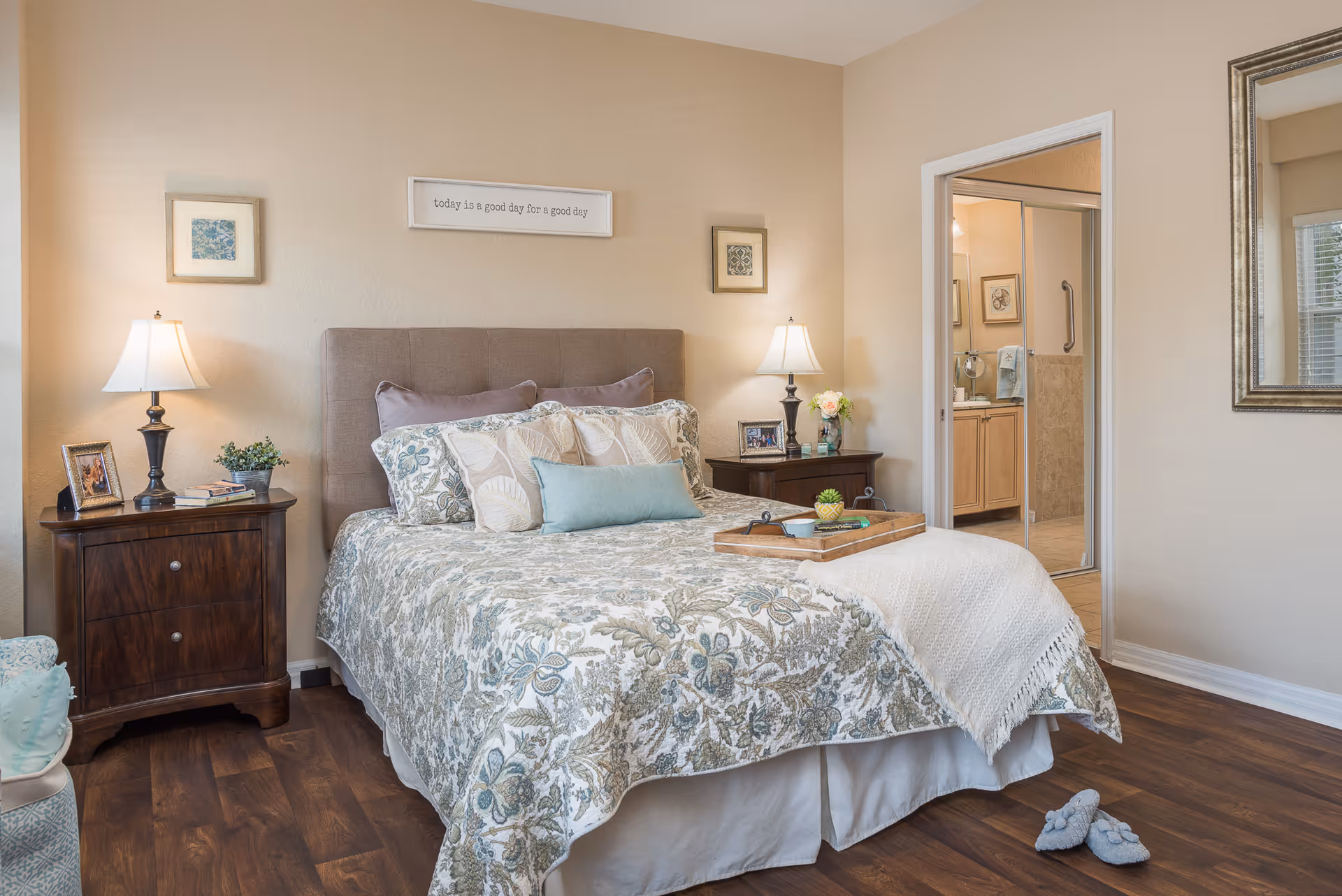 A cozy bedroom with a neatly made bed featuring floral patterned bedding and multiple pillows. There are two wooden nightstands on either side of the bed, each with a lamp and decorative items. A framed sign above the bed reads 'today is a good day for a good day.' The room has wooden flooring and a doorway leading to a bathroom with a glass shower door. A pair of slippers is placed on the floor near the bed.