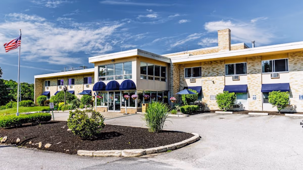 Exterior view of Briarcliffe Manor Skilled Nursing & Rehabilitation building with a two-story structure, beige brick walls, blue awnings over windows and entrance, landscaped bushes and plants, an American flag on a flagpole, and a partly cloudy blue sky.