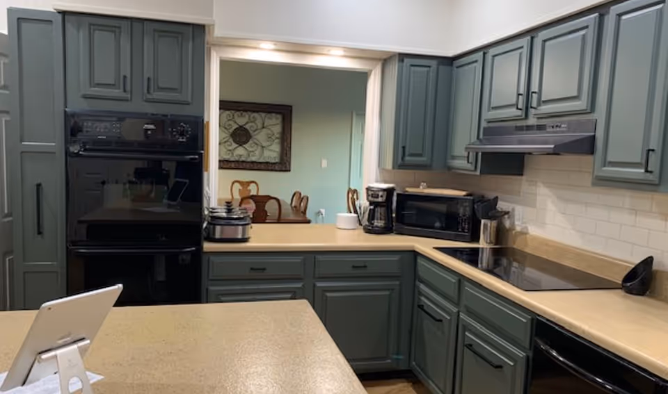 A kitchen with green cabinets and beige countertops. The kitchen features a black built-in oven, a microwave, a coffee maker, a slow cooker, and an electric stovetop. There is a pass-through window opening to a dining room with wooden chairs and a decorative wall hanging.