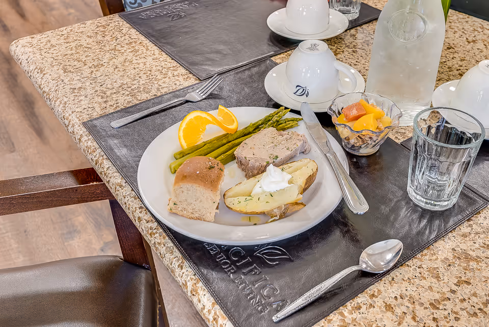 A dining table set with a plate of food including a bread roll, baked potato with sour cream, asparagus, a slice of meatloaf, and orange slices. There is also a small bowl of mixed fruit, a glass of water, a water pitcher, and white cups on saucers on a black placemat.