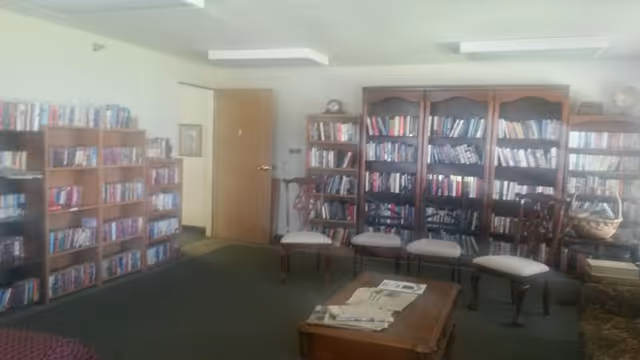 Interior view of a cozy library or reading room with multiple wooden bookshelves filled with books along the walls. There are four wooden chairs with cushioned seats arranged in front of the bookshelves and a wooden coffee table with newspapers on it in the center of the room. The room has a carpeted floor and a door slightly ajar leading to another area.