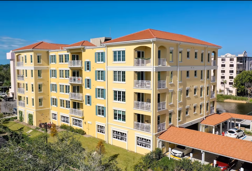 A multi-story yellow building with red-tiled roofs and multiple balconies, surrounded by greenery and a parking area with covered carports. The sky is clear and blue.