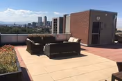 Outdoor rooftop patio area with cushioned seating arranged around a small table. There are planters with greenery on the left side and a view of a city skyline in the background under a partly cloudy sky.