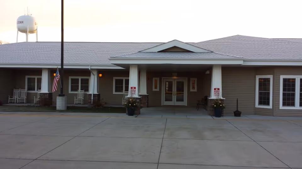 Front exterior view of a single-story assisted living facility building with a covered entrance supported by white columns. There are several windows, rocking chairs on a porch to the left, an American flag, and a water tower visible in the background.
