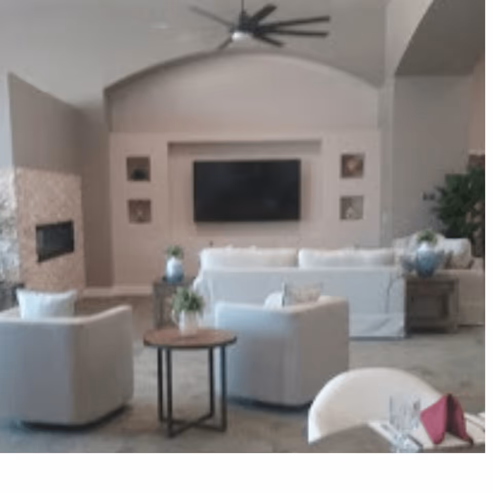 A modern living room with white sofas and armchairs arranged around a small round wooden table with a plant. A large flat-screen TV is mounted on the wall above a built-in fireplace. The room features neutral tones, a ceiling fan, and decorative plants on side tables.