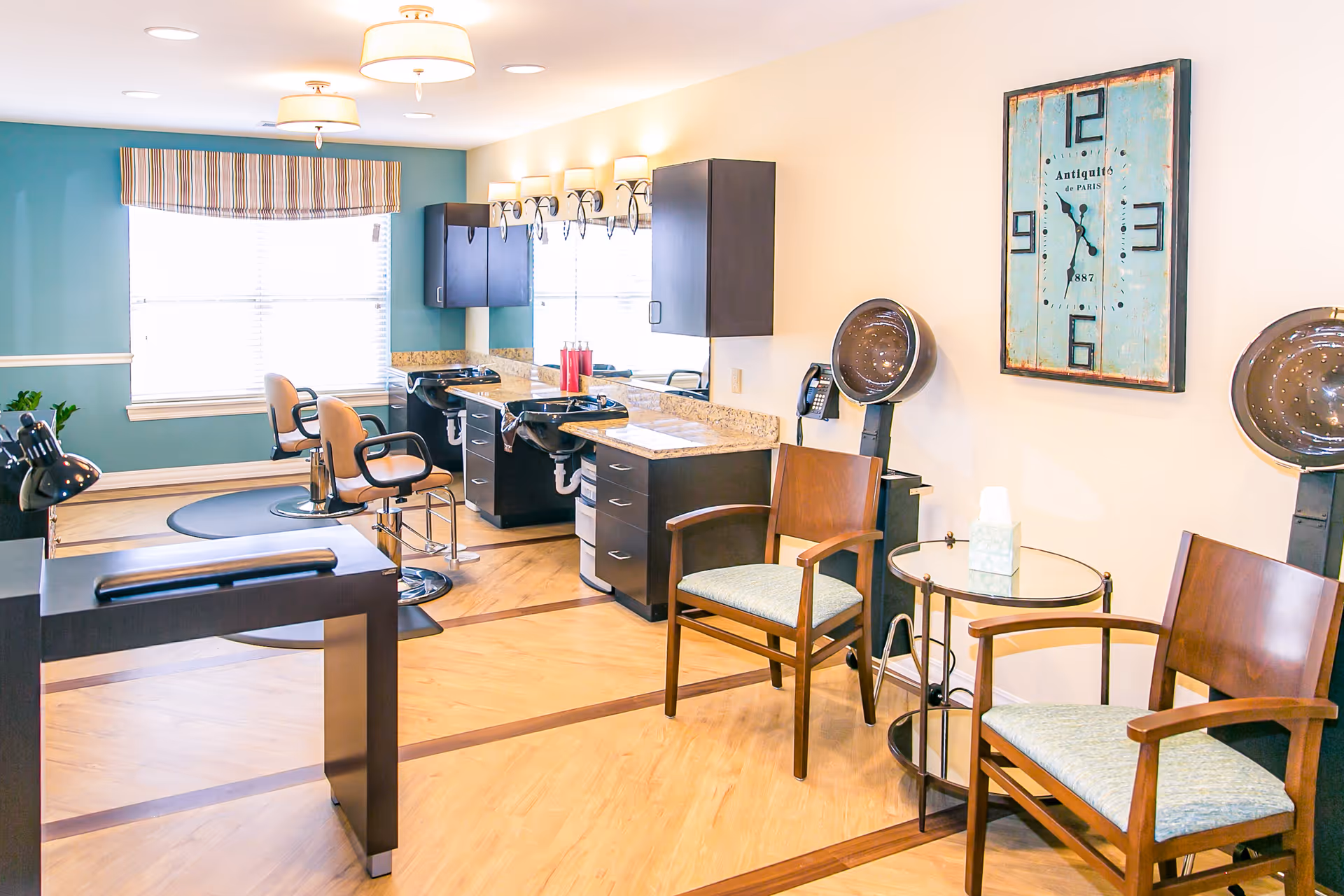 A bright and clean salon area in a senior living facility with two hair washing stations, two salon chairs, two hair dryers, a small round table with a tissue box, and two wooden chairs with cushions. The walls are painted light blue and beige, with a large window covered by a striped valance. A vintage-style wall clock is mounted on the beige wall.