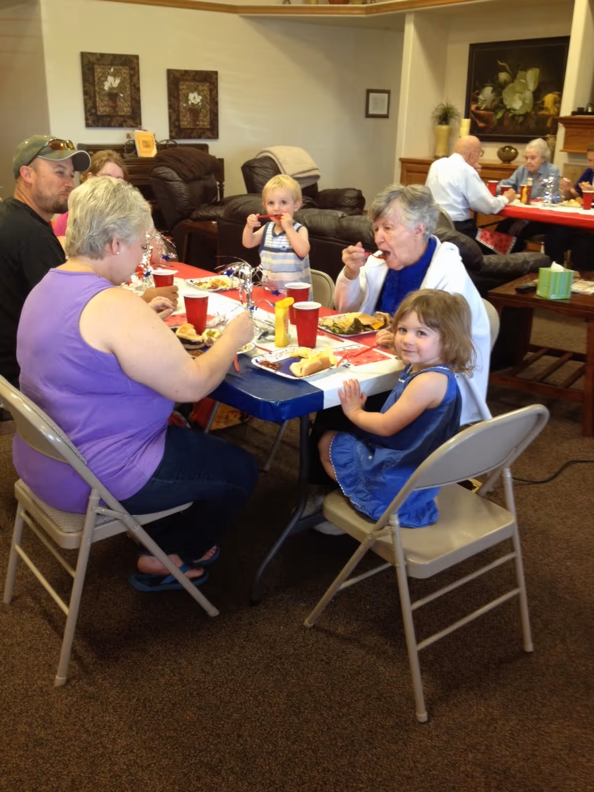 A group of people, including elderly adults and young children, sitting around tables in a common area eating a meal together. The room has comfortable chairs, framed artwork on the walls, and a warm, home-like atmosphere.