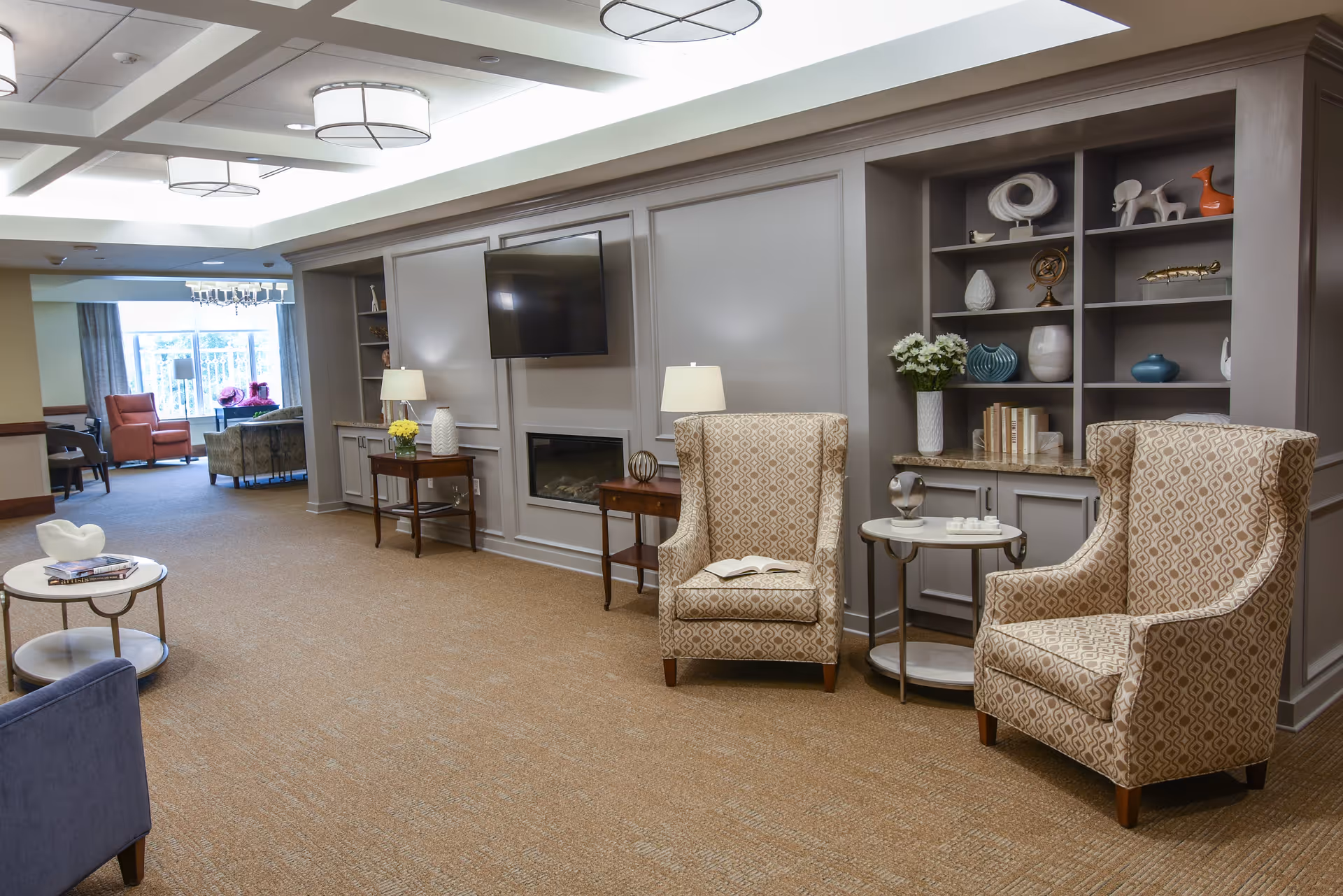 A cozy senior living facility common area with two patterned armchairs, a small round side table, built-in shelves with decorative items, a wall-mounted TV above a fireplace, and additional seating visible in the background near large windows.