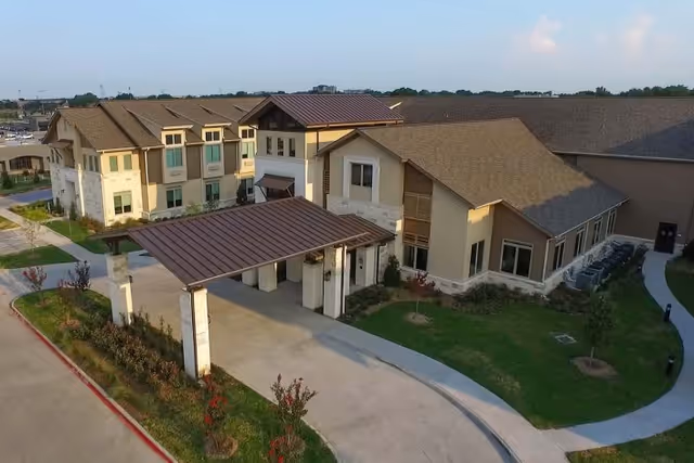 Aerial view of a senior living facility building with a covered entrance driveway, landscaped greenery, and a curved sidewalk. The building has multiple sections with beige and brown exterior walls and a mix of stone and siding materials under a brown shingled roof.