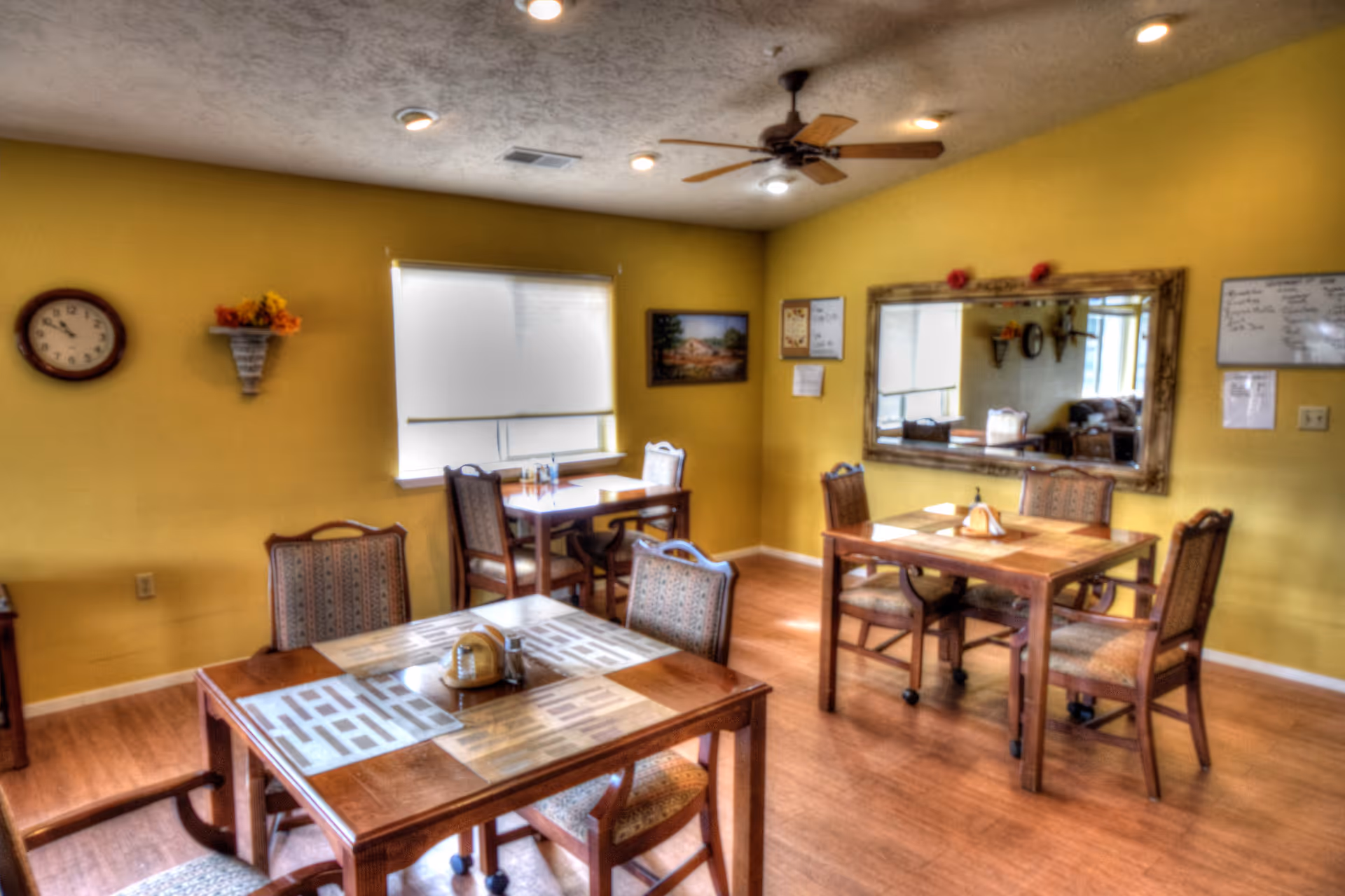 A dining room with yellow walls and wooden flooring featuring three wooden tables each surrounded by four cushioned chairs. The tables have placemats and condiment holders. A large mirror hangs on one wall, reflecting part of the room. There is a ceiling fan with lights, a clock on the wall, a window with a blind, and some wall decorations including a small shelf with flowers and framed pictures.