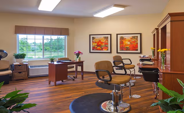 Bright, wood-floored interior salon with styling chairs, workstations, a desk by a window, and framed artwork on the walls.